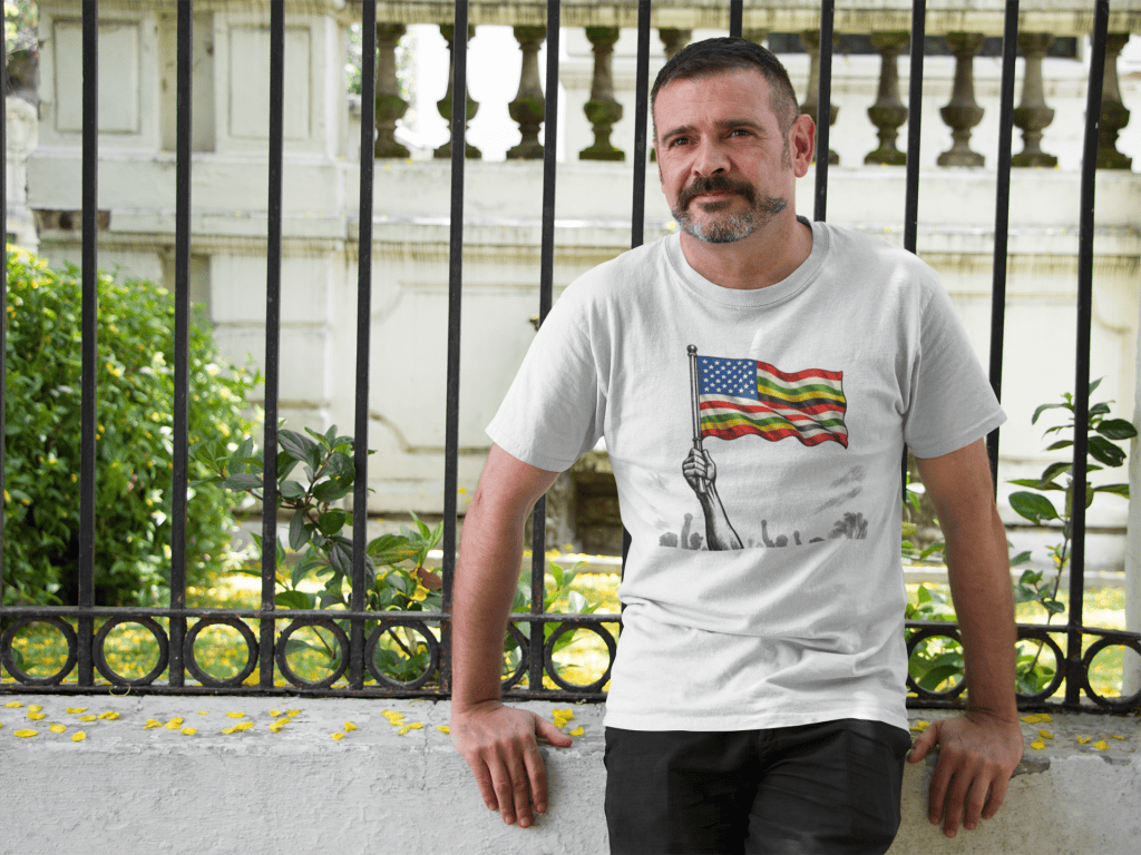 Man in patriotic T-shirt with American flag design outdoors, standing near a decorative fence.