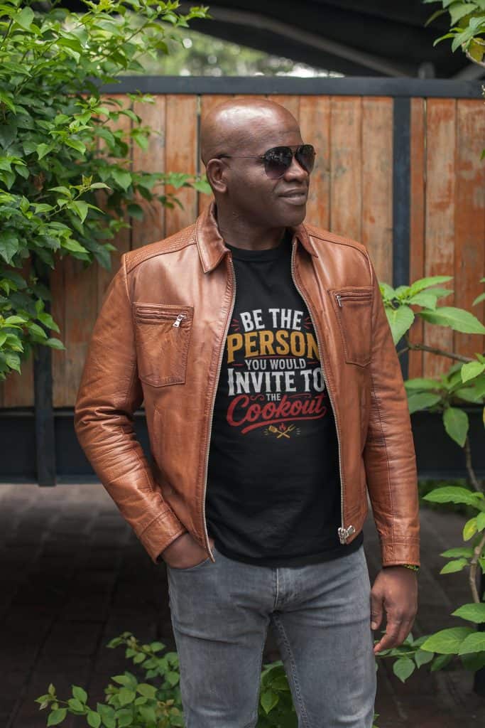 A man wearing sunglasses and a brown leather jacket standing outdoors with greenery and a wooden fen.